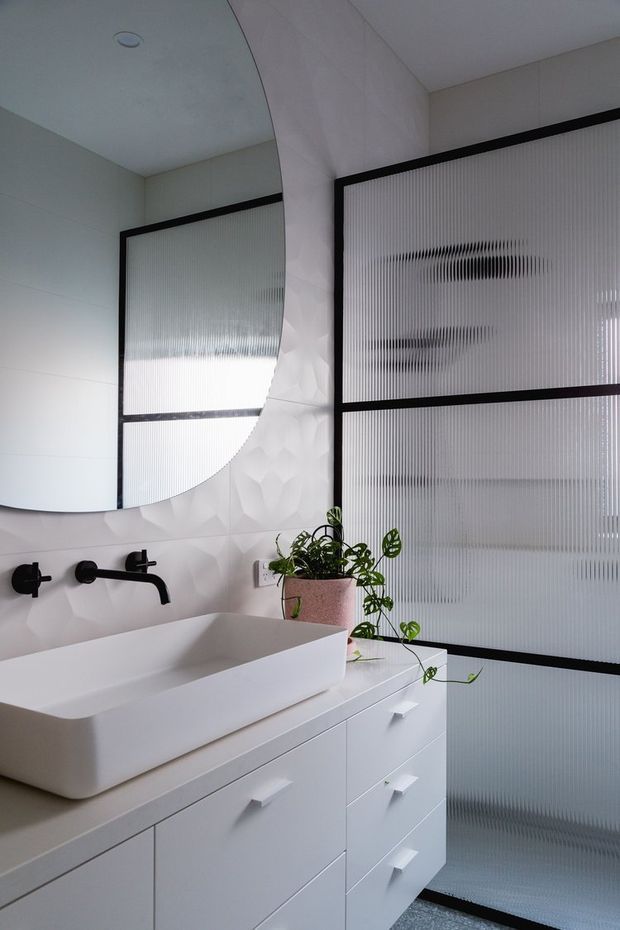 A contemporary bathroom with a white vessel basin set on a white built-in vanity, matte black wall-mounted tapware, and a large rounded mirror. Fluted translucent glazing in black frames fills the background, while a faceted geometric white wall surface and a trailing potted plant add texture and contrast.