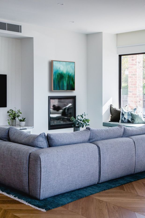 Bright contemporary living room viewed from behind a large blue-grey sectional sofa. The room has white walls and ceiling, a black-framed fireplace, built-in vertical wall detailing, recessed lighting, a large window seat with cushions, abstract artwork, indoor plants, and a dark teal rug over a warm timber parquet floor.