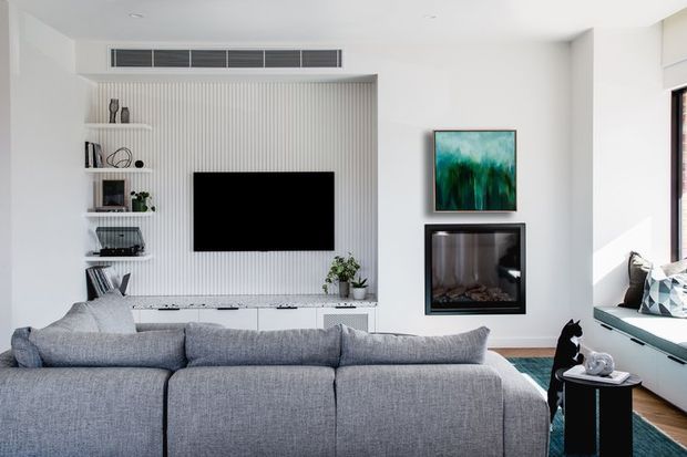 Bright contemporary living room viewed from behind a large grey sectional sofa. The room features a wall-mounted television within a white vertical-ribbed feature wall, integrated white cabinetry and open shelving, a second black-framed fireplace or display unit, artwork, and a built-in window seat with cushions. The palette is predominantly white, light grey, and natural wood, with strong daylight and clean-lined built-in detailing.