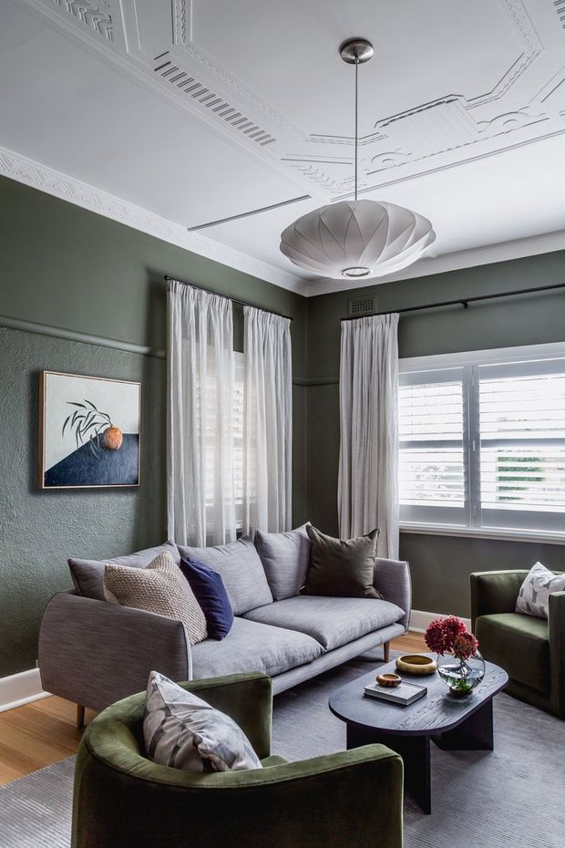 A refined living room with deep muted green walls, a grey upholstered sofa, olive velvet armchairs, and a dark rectangular coffee table. The room features sheer full-length curtains, plantation shutters, timber flooring, and an ornate white decorative ceiling with intricate mouldings and a sculptural pendant light. Layered cushions, artwork, books, bowls, and floral arrangements add a sophisticated, comfortable character.