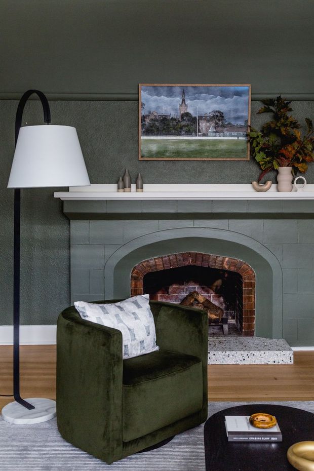 A moody, traditionally styled living room centered on a painted masonry fireplace with a brick-lined firebox and white mantel. The room features dark green textured walls, warm timber flooring, a deep green upholstered armchair, a tall black-and-white floor lamp, a framed landscape artwork, and decorative objects on the mantel. A low dark coffee table and striped rug are partially visible in the foreground.