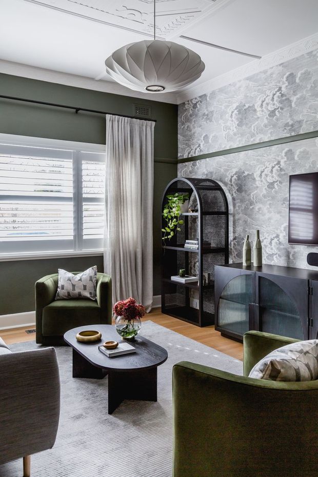 A refined living room with deep olive-green walls, ornate ceiling mouldings, patterned grey wallpaper, plantation shutters, and layered curtains. The room features a sculptural pendant light, curved black open shelving, dark fluted cabinetry, several green upholstered lounge chairs, a rounded black coffee table, and a pale textured rug. The overall look is moody, elegant, and richly layered.
