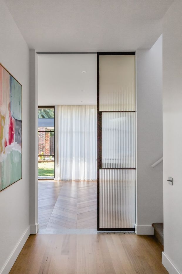 Bright contemporary interior view from a hallway or entry area toward an open living space. The scene features pale timber flooring, white walls and ceiling, a large abstract artwork, sheer curtains, a garden-facing glazed opening, and a striking black-framed ribbed glass partition or door. A stair edge is visible at right.