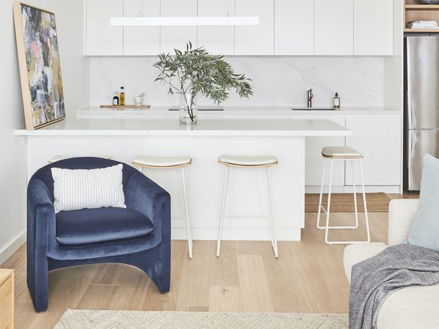 Bright open-plan kitchen and living area with white flat-panel cabinetry, a pale stone island and splashback, light timber flooring, and three bar stools. A navy upholstered armchair and cream sofa frame the foreground, with a large vase of leafy branches on the island and a framed artwork leaning against the wall. The space has a clean, contemporary aesthetic with abundant natural light.