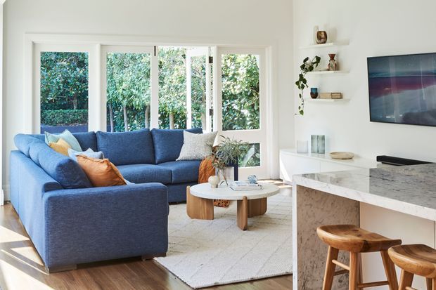 Bright contemporary open-plan living and kitchen area with a deep blue sectional sofa, neutral rug, white walls, large garden-facing windows, floating shelves, wall-mounted television, and a marble-look kitchen island with timber stools.