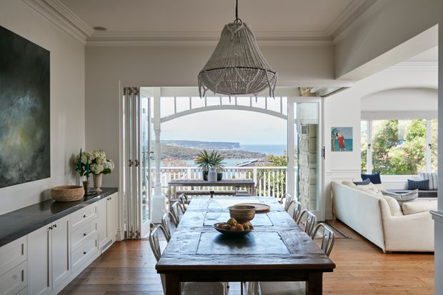A refined open-plan dining room overlooking a coastal harbour or bay. The room features a long rustic timber dining table with metal chairs, white built-in cabinetry, decorative wall panelling and cornices, polished timber flooring, and a large woven pendant light. Open glazed doors lead to a balcony or terrace, with a bright living room visible to the right and a scenic waterfront landscape beyond.