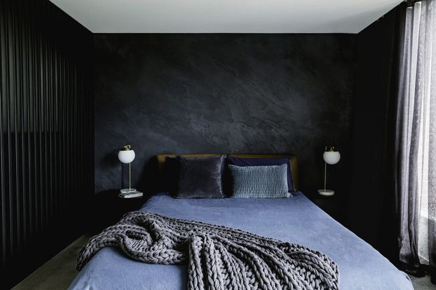 Moody contemporary bedroom with a dark textured feature wall, vertically ribbed black wall panelling, a neatly made bed in muted blue-grey bedding, layered pillows, and a chunky knitted throw. Minimal bedside lamps and full-height curtains frame the room.