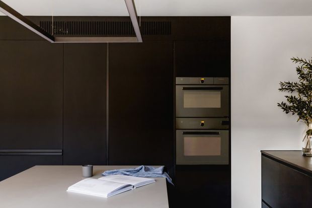A refined contemporary kitchen with tall matte dark cabinetry, integrated double wall ovens, a pale stone island or worktop, and a minimalist black rangehood. A white wall and a small plant add contrast, while an open book and cloth rest on the island.