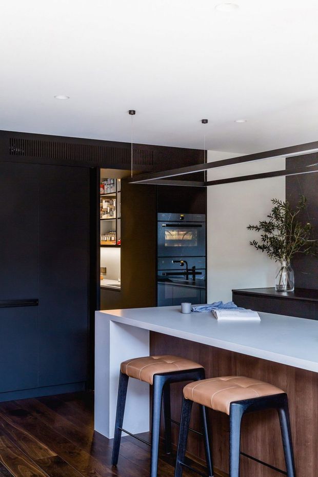 A contemporary kitchen featuring a large white island with a dark timber-clad base, two tan leather bar stools, integrated black appliances, dark cabinetry, open pantry shelving, and a dark timber floor. A minimalist vase with foliage sits on a dark sideboard, while slim linear pendant lights hang above the island.