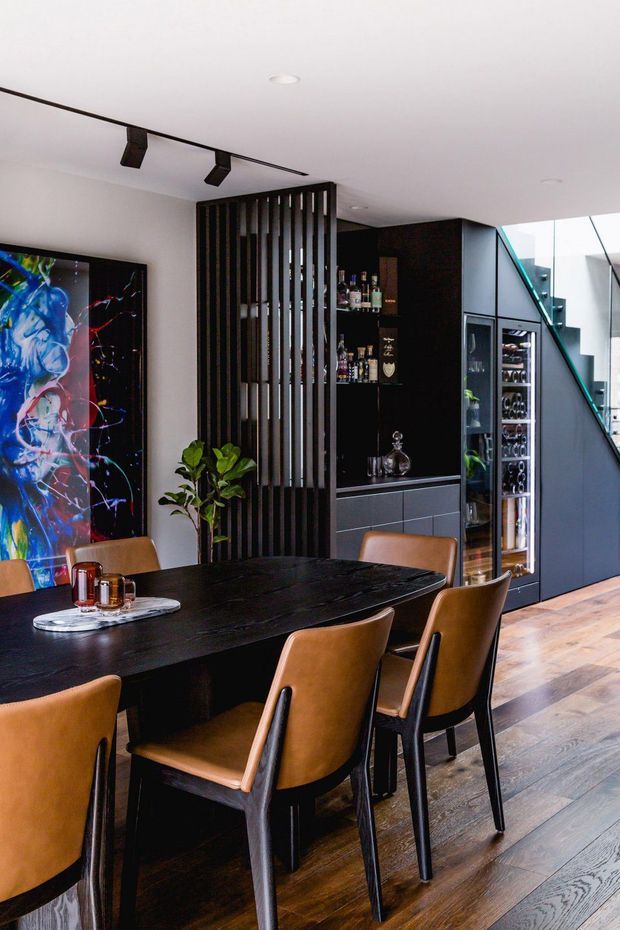A contemporary dining area with a long black dining table and brown leather dining chairs. The room features dark timber-look flooring, black built-in cabinetry with an integrated drinks bar and glass-front storage, and a vertical black slatted room divider. A colourful abstract artwork and indoor plant add visual accents, while a glazed stairwell brings in natural light.