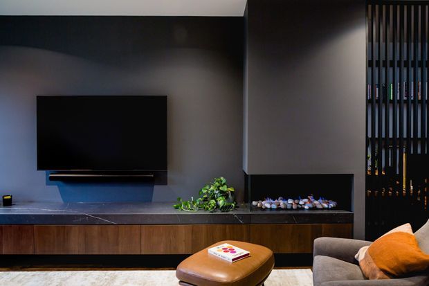 A dark, refined contemporary living room featuring a wall-mounted flat-screen TV above a long built-in timber media cabinet with a dark stone benchtop. A low suspended blue-grey feature panel sits beneath the TV, with decorative plants and stones on the cabinet. The room includes a brown leather ottoman, a grey sofa with an orange cushion, and a vertical timber privacy screen at the right.