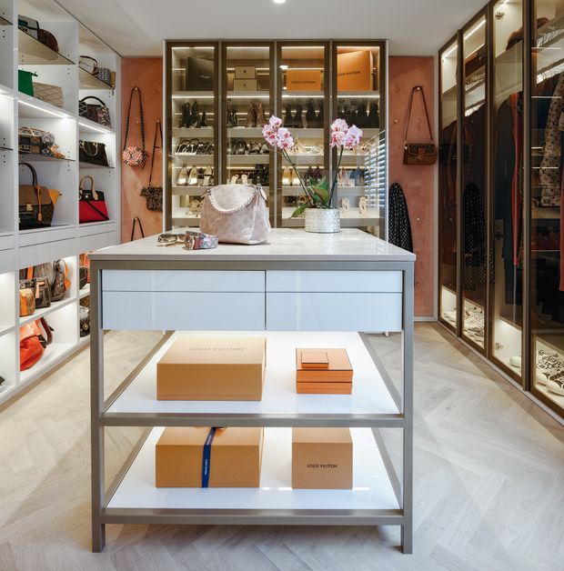 A luxurious walk-in wardrobe and dressing room centered on a large island display unit. The space features extensive illuminated glass-front cabinetry and open shelving filled with handbags, shoes, clothing, and accessories. Warm wood-toned wall panels, bronze or dark metal-framed wardrobes, pale cabinetry, and light herringbone timber flooring create a refined boutique-like interior. A central white stone-topped island provides drawers and open display shelves, while integrated lighting highlights the contents.