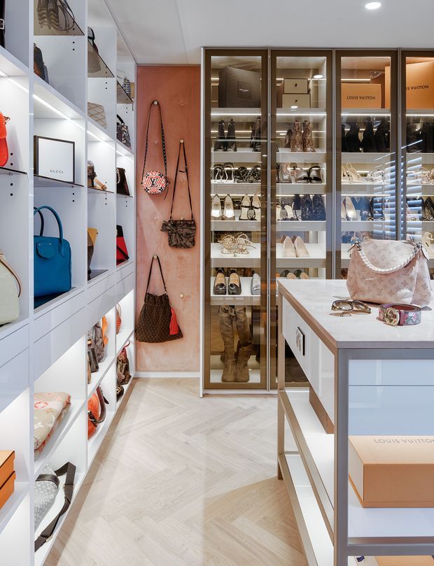 A bright, highly organized luxury walk-in wardrobe and dressing room displaying handbags, shoes, clothing, and accessories in illuminated open shelving and glass-front cabinetry. A central island with a pale stone benchtop provides additional storage and display space. The room features pale herringbone timber flooring, white joinery, warm wood-toned backing panels, bronze-toned cabinet frames, and integrated LED lighting.