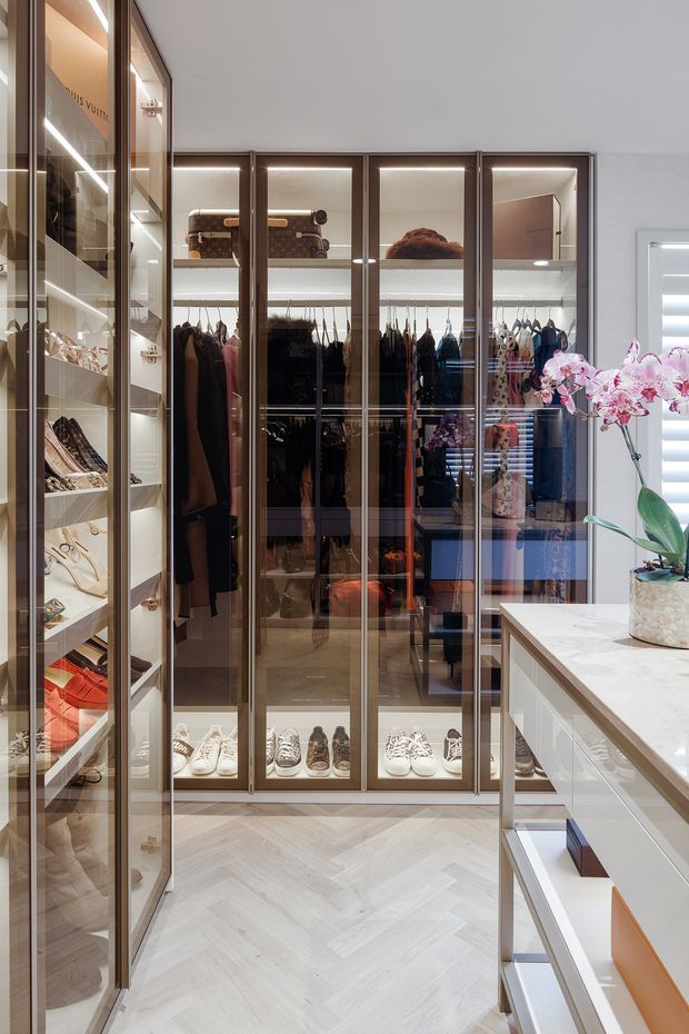 A luxurious walk-in wardrobe with floor-to-ceiling bronze-framed glass cabinetry, illuminated shelving, hanging rails, shoe storage, and display compartments. A central white stone-topped island with drawers occupies the right foreground, while a potted orchid adds a decorative touch. The space has a light herringbone floor, warm integrated lighting, and highly organised built-in storage.