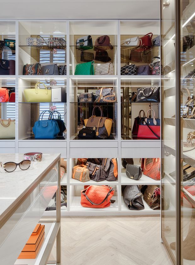 A brightly lit luxury walk-in wardrobe or boutique dressing room with floor-to-ceiling white built-in shelving and glass-fronted display cabinets filled with handbags. A marble-topped island or display table sits in the foreground with sunglasses and accessories, while mirrored panels reflect the cabinetry. The room has pale herringbone flooring, integrated shelf lighting, and a refined retail-like presentation.