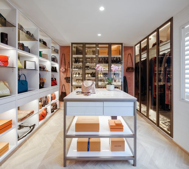 A bright, highly organised walk-in wardrobe and dressing room with extensive open shelving, illuminated glass-front cabinetry, hanging rails, handbags, shoes and accessories. A central island with a pale stone benchtop and open display shelves anchors the room. The space has warm timber-framed cabinetry, white joinery, herringbone flooring, recessed ceiling lights and a window with plantation shutters.