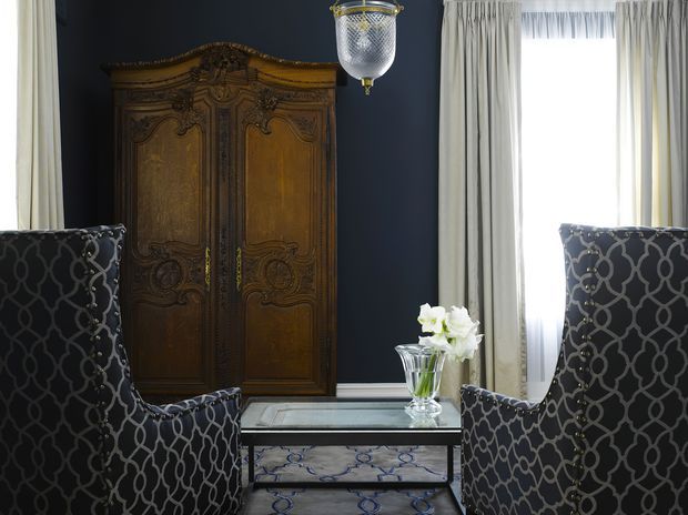 A refined living room vignette featuring two tall patterned armchairs facing a mirrored coffee table, an ornate dark wood armoire against a deep charcoal wall, floor-length cream curtains, a patterned rug, and a decorative hanging lantern. The room has a formal, luxurious traditional character with strong contrast between dark upholstery and light drapery.