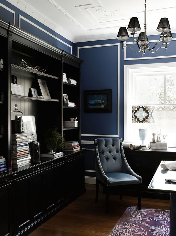 A refined home office with deep navy-blue walls framed by white decorative mouldings, a large black built-in bookcase, dark timber flooring, and a tufted charcoal leather desk chair. A desk sits beside a bright window, with a traditional multi-arm chandelier overhead and curated books and objects displayed in the joinery. The room has a formal, richly layered, classic character.