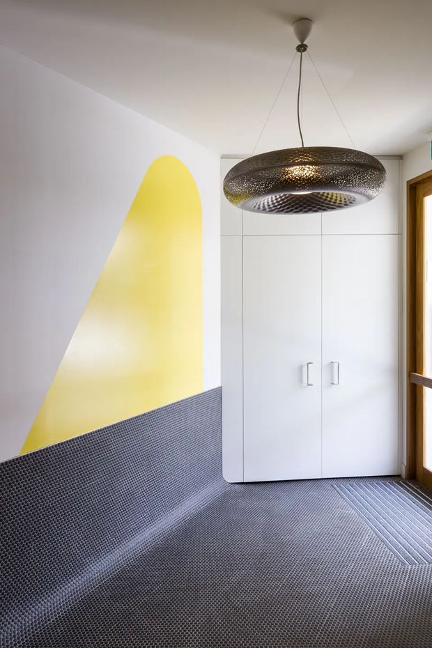 A compact, highly stylised entryway or hall with a white built-in storage cupboard, dark textured patterned flooring and matching lower wall treatment, a bold yellow geometric wall feature, and a large perforated sculptural pendant light. Timber-framed glazing or a door is visible at the right edge. The space has a contemporary, graphic interior with strong contrast and distinctive custom finishes.