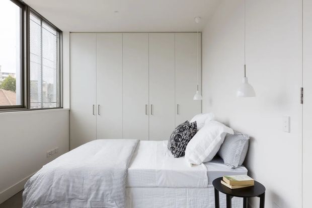 Bright, compact minimalist bedroom with a neatly made bed, white built-in floor-to-ceiling wardrobes, large framed windows with blinds, and simple pendant lights. The room uses a restrained white and grey palette with subtle patterned bedding and a small black bedside table.