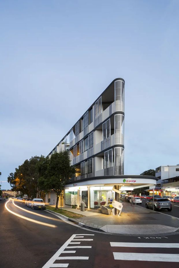 Street-level exterior photograph of a contemporary multi-storey mixed-use building on a corner site. The building has a narrow, curved glazed corner, extensive floor-to-ceiling windows, dark metal framing, and ground-floor commercial frontage occupied by a St.George bank. Urban streets, vehicles, signage and neighbouring buildings surround the property.