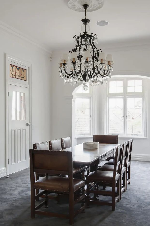 Bright formal dining room with white walls, ornate ceiling moulding and medallion, a large black crystal chandelier, tall multi-pane bay windows, decorative glazed door panel, and a dark timber dining table with upholstered leather chairs. The room combines traditional architectural detailing with refined period-inspired furnishings.