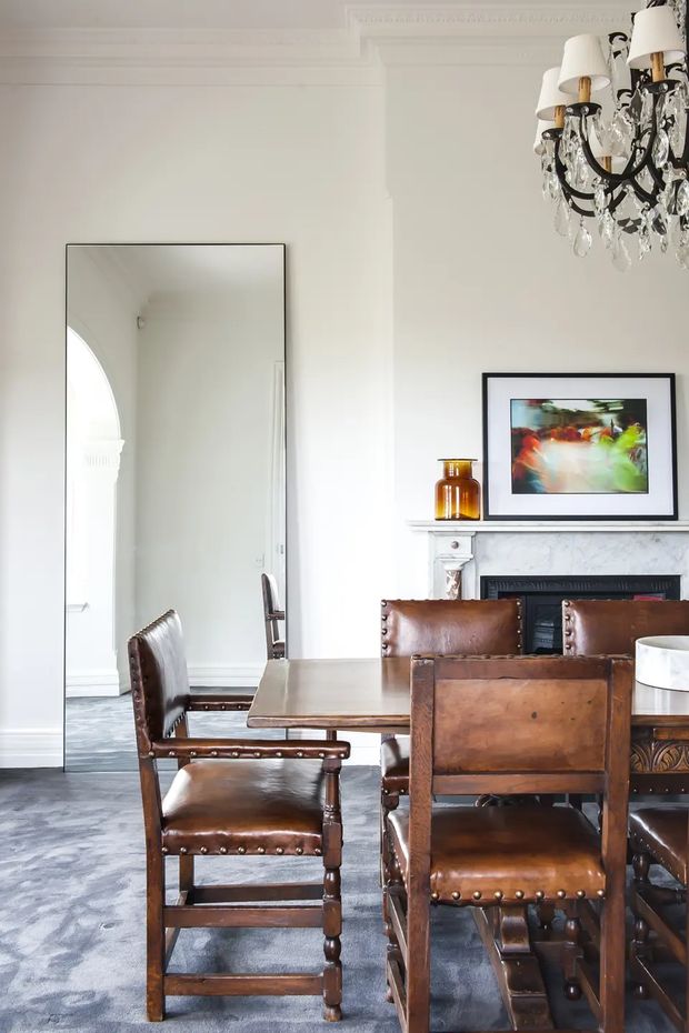 Bright formal dining room with a substantial dark timber dining table and matching leather-upholstered, nailhead-trimmed chairs. The room features white walls with ornate traditional cornice detailing, a large wall mirror, a decorative fireplace with white mantel, framed artwork, and an elaborate black crystal chandelier. Grey patterned carpet and rich brown leather and wood create a refined classic interior.