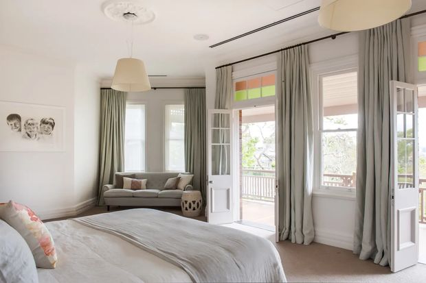 Bright, spacious bedroom with a large upholstered bed in the foreground, pale neutral bedding, grey curtains, and a small sofa beneath tall windows. French doors open onto a balcony or veranda, with colourful transom glazing and detailed ceiling roses adding character. The room combines traditional architectural detailing with restrained contemporary furnishings.