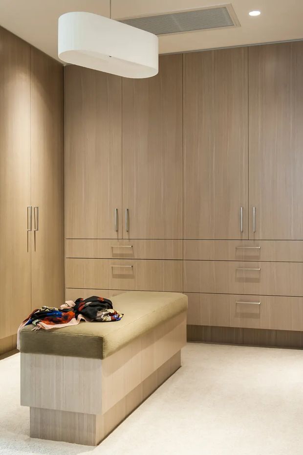 A refined walk-in wardrobe or dressing room with extensive floor-to-ceiling light wood cabinetry, integrated drawers and slim metal handles. A central upholstered island provides seating and storage, while a large white pendant light and recessed ceiling lighting illuminate the warm, neutral space. The room has a clean, contemporary built-in design with pale carpet flooring.
