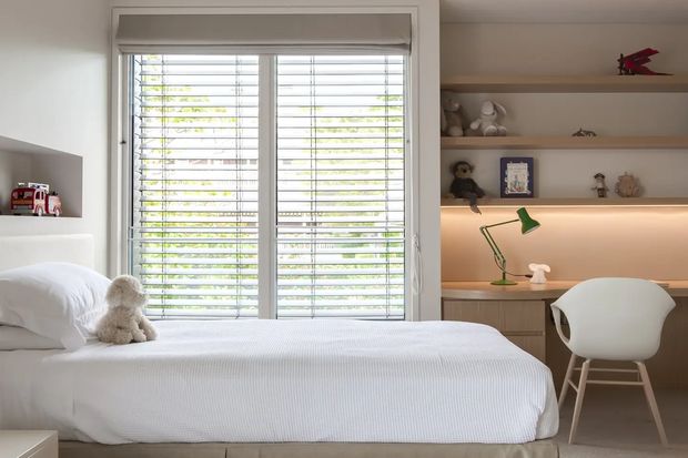 Bright, tidy children's bedroom with a white upholstered bed, soft bedding and a teddy bear, built-in open shelving, and a compact study desk beneath warm integrated shelf lighting. A large window with white plantation shutters provides abundant natural light, while neutral finishes and a small green desk lamp add gentle colour.