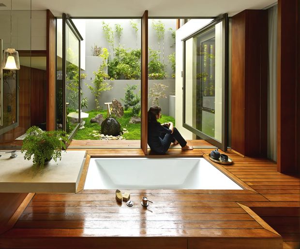 A serene Japanese-inspired bathing space with a sunken white soaking tub set into a warm timber floor, opening through large glazed doors to a private planted courtyard. Rich dark timber wall and door frames, indoor greenery, and integrated low-level joinery create a calm connection between interior and garden.