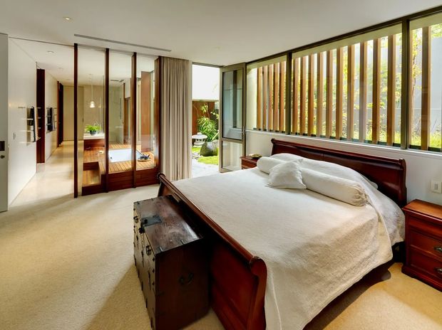 A bright, spacious bedroom with a large dark timber bed, white bedding, matching bedside furniture, and a vintage timber trunk at the foot of the bed. Floor-to-ceiling glazing with vertical timber sun-shading screens runs along one wall, overlooking greenery. The room connects visually to a tiled bathroom or dressing area through glazed timber-framed partitions. Neutral carpet, white walls, and warm timber details create a calm, refined interior.