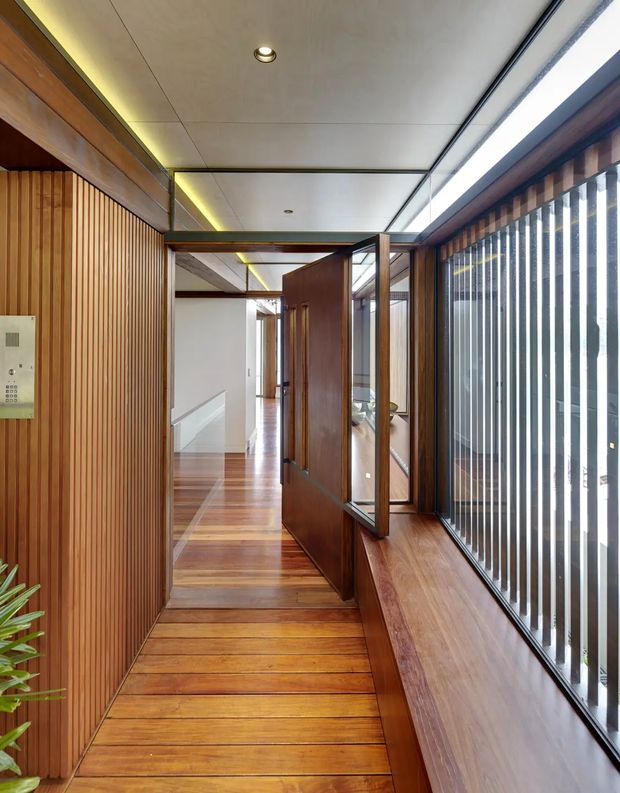 A narrow, warm-toned interior passage or landing with rich timber flooring, vertical timber wall battens, timber-framed glazing, and a long built-in timber window seat. A dark timber door opens toward a brighter hallway, while vertical external screening filters daylight along the right side. The ceiling is composed of clean pale panels with recessed downlights and concealed linear lighting, creating a refined modern architectural interior.