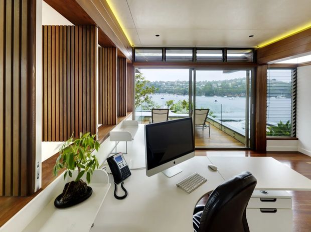 A bright contemporary home office with a built-in white desk and cabinetry, desktop computer, telephone and black leather office chair. Warm timber wall and ceiling detailing includes vertical timber battens and broad timber framing. Floor-to-ceiling glazing opens to a small timber balcony with outdoor chairs and clear views across a waterfront inlet and surrounding homes. Concealed linear lighting adds a warm glow along the ceiling perimeter.