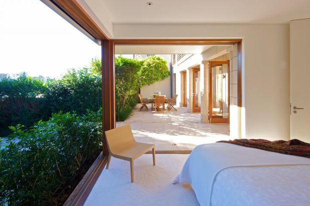 Bright bedroom opening through large timber-framed glazing and wide sliding doors onto a sunny paved courtyard. The room contains a partially visible bed and simple light-coloured chair, while the courtyard beyond has timber-framed doors, outdoor dining furniture, and lush screening vegetation. The design is clean and contemporary with strong indoor–outdoor connection and abundant natural light.