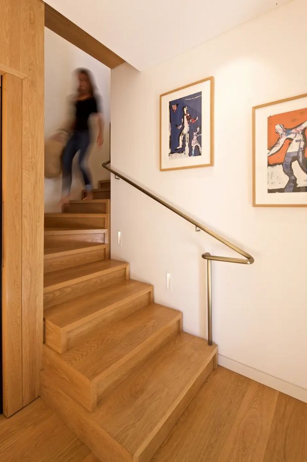 Interior view of a warm, compact timber stairway with broad oak-toned stairs and matching wood flooring. A slim bronze or dark metal handrail follows the white-painted stair wall, while a timber-framed opening and vertical joinery frame the stairwell. Two framed artworks decorate the wall, and a blurred person is moving on the upper stairs. The space has clean contemporary detailing and warm natural materials.