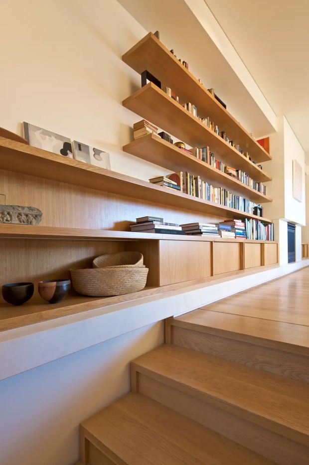 Warm contemporary interior view of a timber-lined stairway and landing, featuring extensive built-in oak-toned shelving, cabinetry, books, and display objects along a white wall. Broad timber steps rise to the landing, with integrated low storage and open shelves creating a library-like feature. The space is brightly lit with a calm, refined, natural palette.