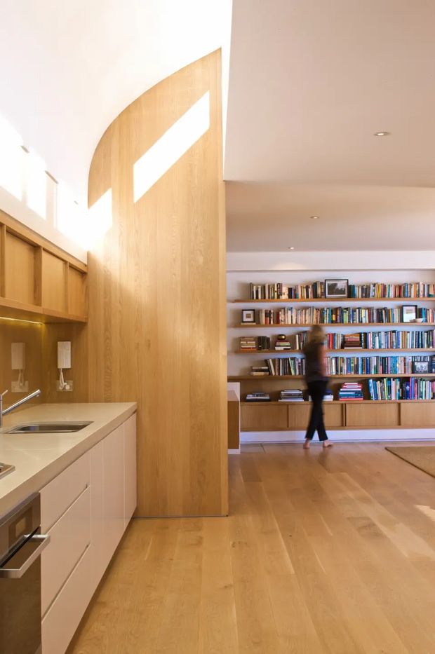Bright open-plan interior showing a minimalist kitchen in the foreground and a living/library area beyond. The kitchen has pale wood cabinetry, a light stone benchtop, integrated appliances and a tall curved timber partition or feature wall. The adjoining space features extensive built-in wall-to-wall bookshelves, timber flooring and recessed ceiling lights. Strong daylight creates a warm, airy atmosphere.