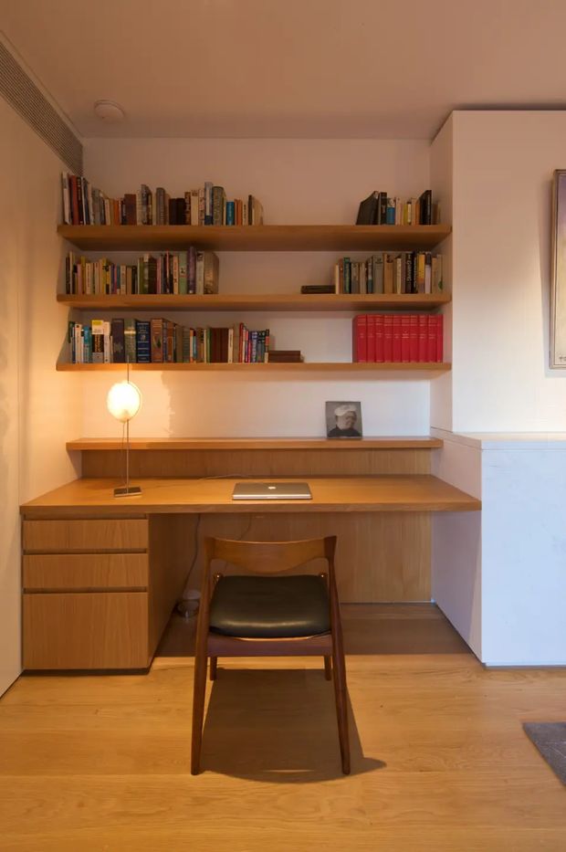 A warm, compact home office nook with a built-in timber desk, drawer storage, floating bookshelves, and a mid-century-style wooden chair. A laptop sits on the desk, while a pendant lamp provides focused ambient lighting. The space has white walls, timber flooring, and abundant books, creating a cozy study atmosphere.