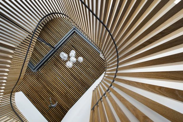 Upward-looking architectural interior view of a sculptural stairwell or circulation void. Curved black metal stair rails and a suspended landing frame a dramatic ceiling and wall treatment of closely spaced timber slats, creating a radial, flowing pattern around a central contemporary light fitting. White surfaces between the timber battens provide strong contrast.