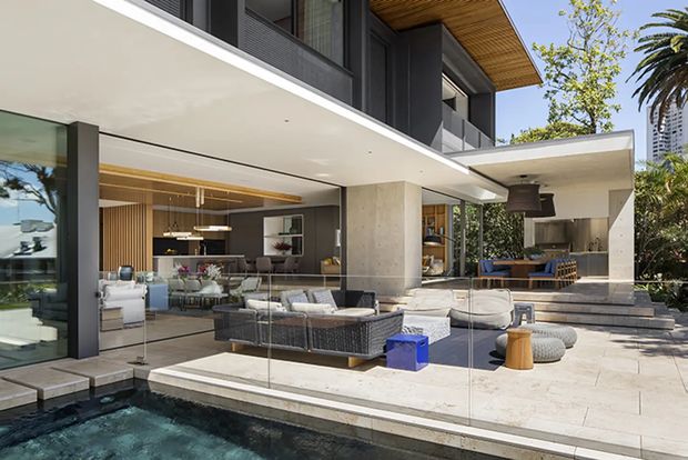 Contemporary multi-level residence with broad cantilevered roof slabs, dark framed glazing, warm timber soffits, and open-plan interiors extending onto a paved poolside terrace. The view centres on the outdoor living area and swimming pool, with a living room, kitchen, and outdoor kitchen/lounge visible behind extensive glazing.