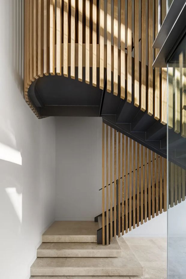 A contemporary interior stairway with pale stone steps, a dark metal stair structure, and a distinctive curved balustrade formed from closely spaced vertical timber slats. White plaster walls and extensive glazing create a bright, refined architectural setting.
