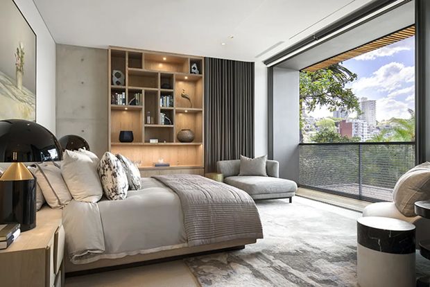 Contemporary bedroom with a large upholstered bed, layered neutral bedding, a built-in timber shelving and storage wall, concrete-look wall panel, and a compact lounge chair. Full-height glazed doors open to a balcony with an urban outlook, bringing in abundant natural light. The room combines warm timber joinery, soft textiles, grey tones, and black metal accents in a refined, modern setting.