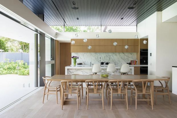 Bright open-plan dining and kitchen interior with a long timber dining table and matching wooden chairs in the foreground. A contemporary kitchen features warm timber cabinetry, a white marble-look splashback, a central island with seating, integrated appliances, and multiple pendant lights. Full-height glazing and a large open doorway connect the room to a leafy outdoor area, while a dark linear ceiling adds contrast.