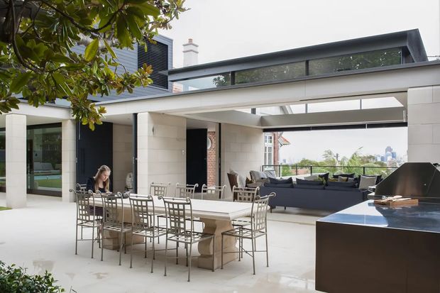 A bright, open-air contemporary entertaining area with a large dining table and metal dining chairs in the foreground, a lounge seating zone beyond, and a dark built-in outdoor kitchen or barbecue island at right. The space is framed by white masonry walls and broad openings, with dark horizontal louvred or screened elements above and views toward a leafy urban setting.