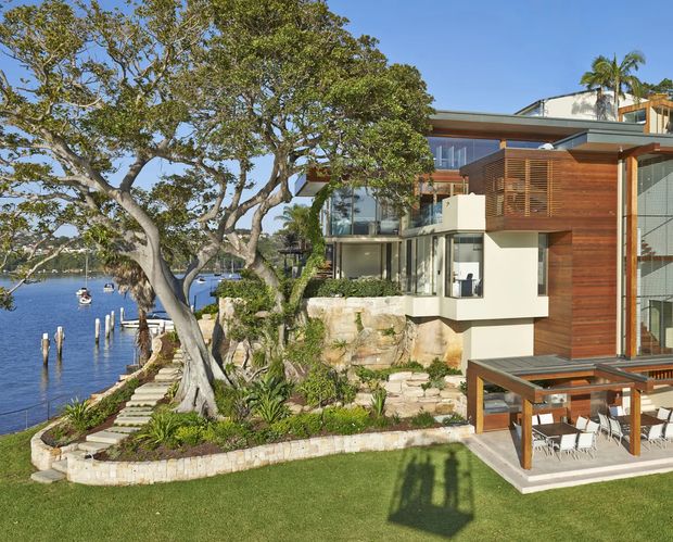 Exterior view of a substantial multi-level waterfront residence set directly beside a calm inlet. The contemporary house combines pale rendered walls, warm timber cladding and screening, extensive glazing, projecting flat roof forms, and a large elevated outdoor dining terrace. Landscaped stone retaining walls, steps, lawn, mature trees, palms, and a small private shoreline with timber jetty posts are visible.