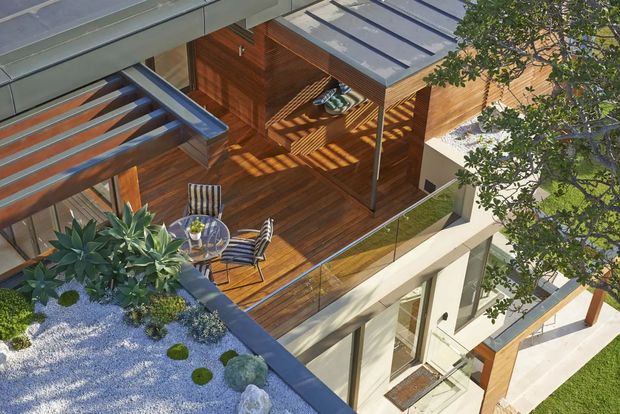 Aerial view of a contemporary multi-level home featuring a spacious timber rooftop terrace, glazed and louvred roof structures, outdoor dining furniture, planted roof areas with succulents and gravel, and crisp white and timber exterior elements. Strong sunlight creates patterned shadows across the deck and walls.