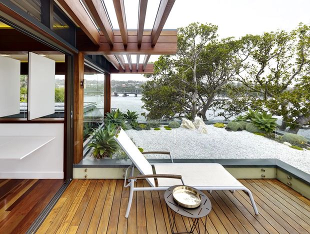 A contemporary waterfront terrace or balcony with warm timber decking, exposed timber pergola beams, and large glazed openings beside a minimalist white interior. A built-in planter bed filled with white gravel and sculptural succulents separates the seating area from the water view. The space includes a white outdoor lounge chair and a low round table, creating a refined indoor-outdoor setting.