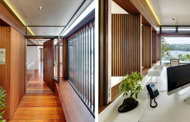 A split-view interior showing a warm timber-lined hallway and a bright contemporary home office. The hallway features polished timber flooring, vertical timber wall slats, timber-framed glazing with vertical screening, recessed ceiling lighting, and a long linear skylight or clerestory. The office includes a white built-in desk, computer monitor, telephone, task chair, indoor plant, and views through glazing toward a leafy waterfront or hillside setting.