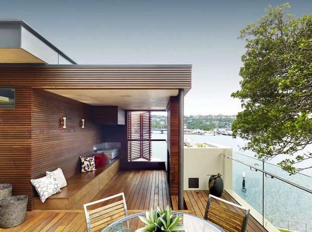 Contemporary timber-clad outdoor terrace or balcony with a covered entertaining area, built-in bench seating, barbecue, timber decking, and glass balustrade overlooking a waterfront or harbour. Horizontal timber slats and louvred screening create a warm, modern architectural character, with outdoor dining furniture in the foreground.